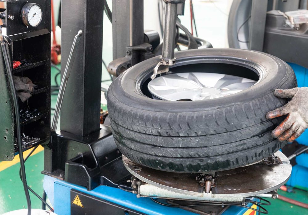 A mechanic is installing a tire onto a wheel using a tire mounting machine in a workshop. The scene shows tools and equipment commonly found in a tire service area.